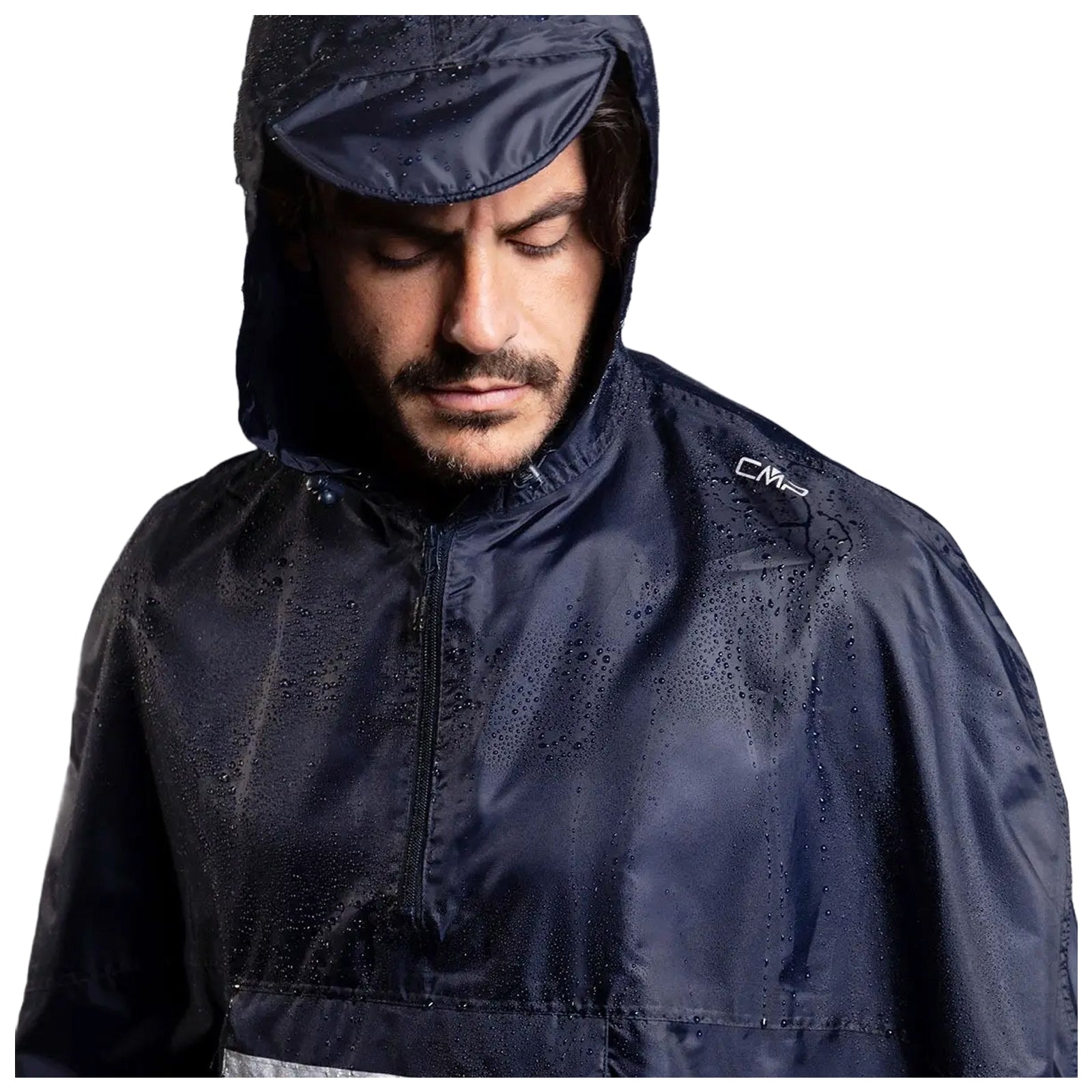 A man wearing a waterproof navy jacket stands still with water droplets on the fabric looking downwards in a neutral expression against a plain background.