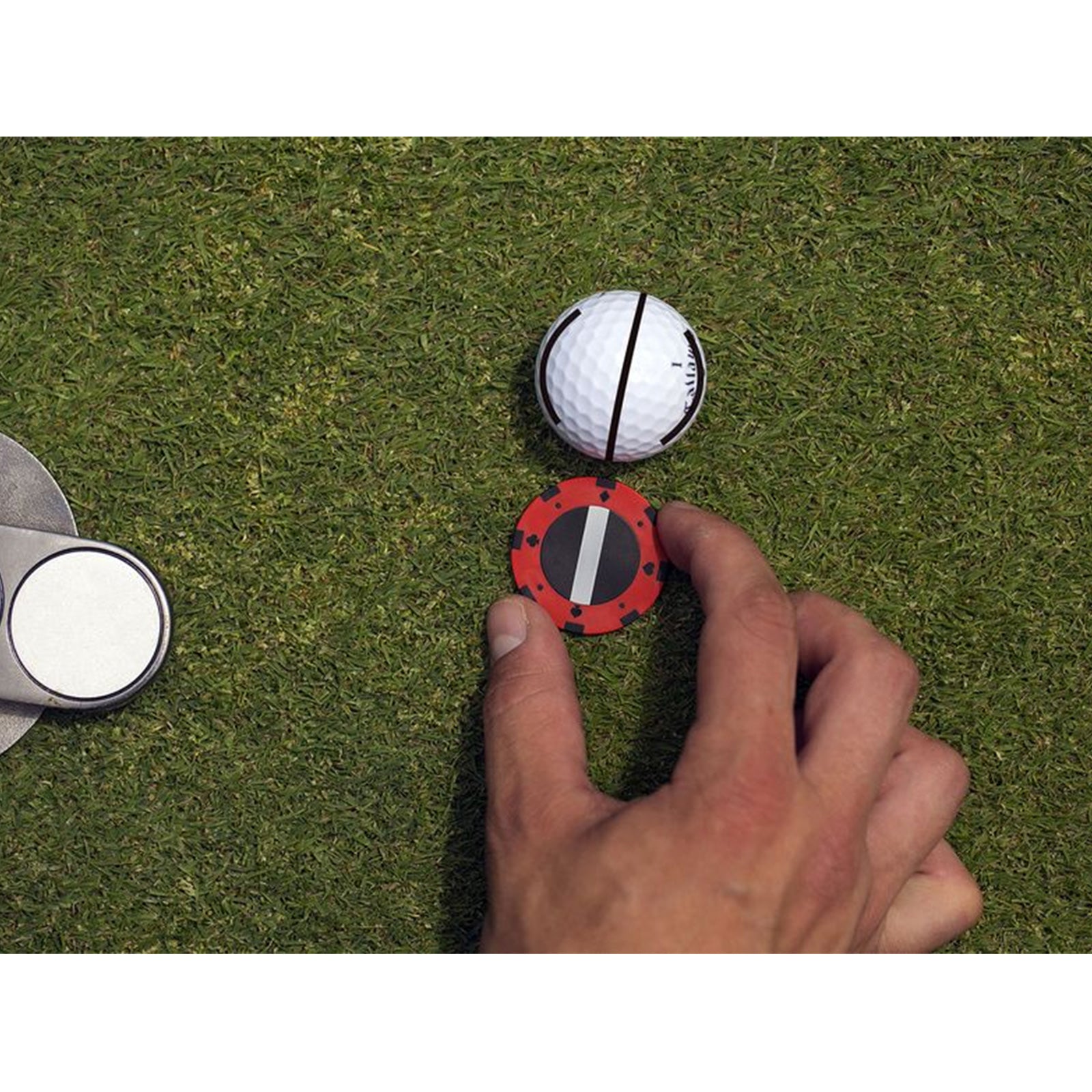 A hand is placing a red marker with a black line on the green grass next to a white golf ball with black stripes while a silver putting device is nearby