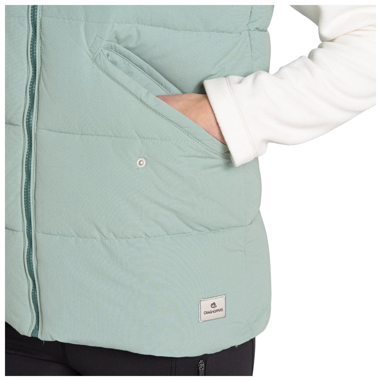 A hand is reaching into a pocket of a light green puffer vest with a white sleeve visible beside it. The vest has a small logo patch at the bottom.