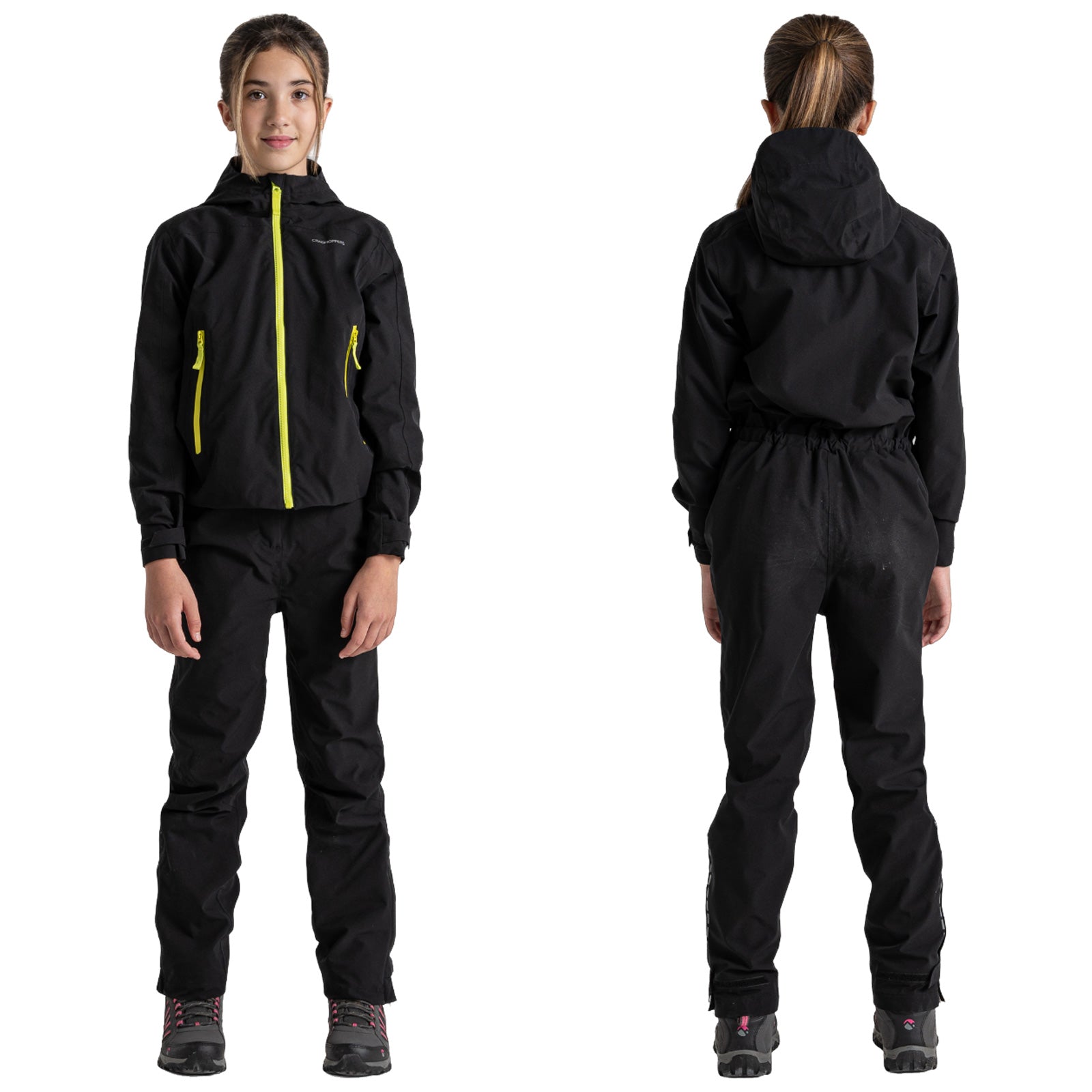 A child is wearing a black waterproof jacket and pants with bright yellow zippers and equipped with hiking boots standing in a neutral studio background.