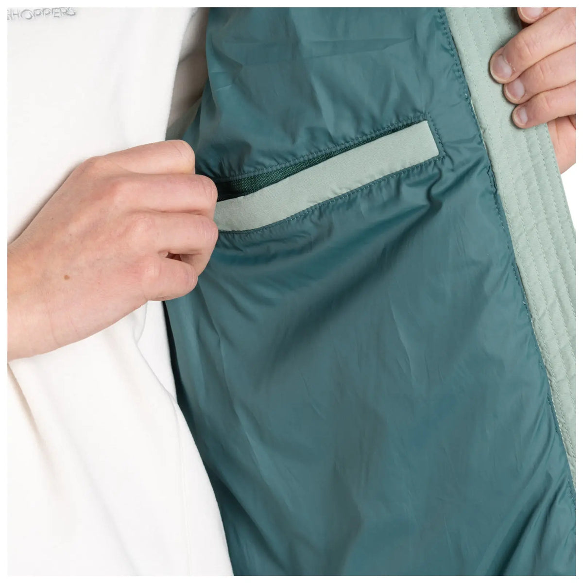 A person’s hand is reaching into a small pocket located inside a green jacket. The surrounding environment is not visible but suggests an indoor setting.