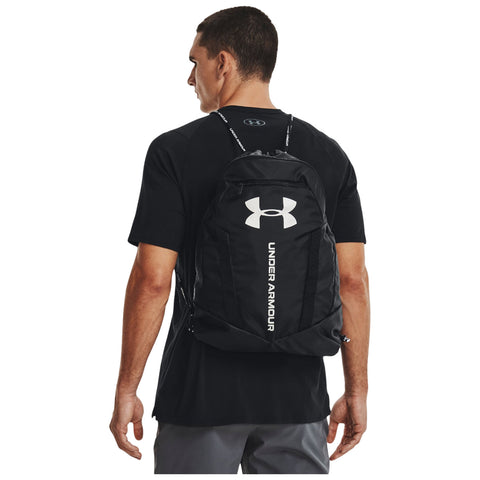 A man wearing a black t-shirt carries a black Under Armour backpack on his back while looking to the side in a neutral setting.