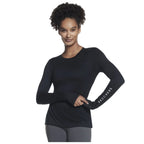 A woman with curly hair is posing confidently while wearing a fitted long-sleeve black top and grey pants with the word SKECHERS printed on her sleeve in white.