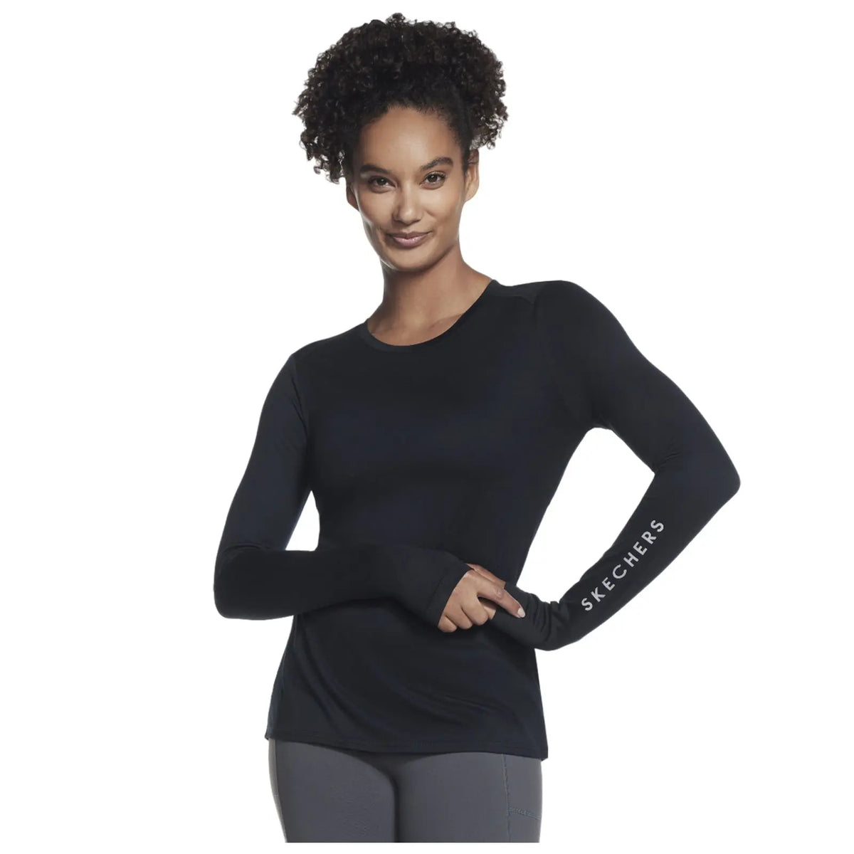 A woman with curly hair is posing confidently while wearing a fitted long-sleeve black top and grey pants with the word SKECHERS printed on her sleeve in white.