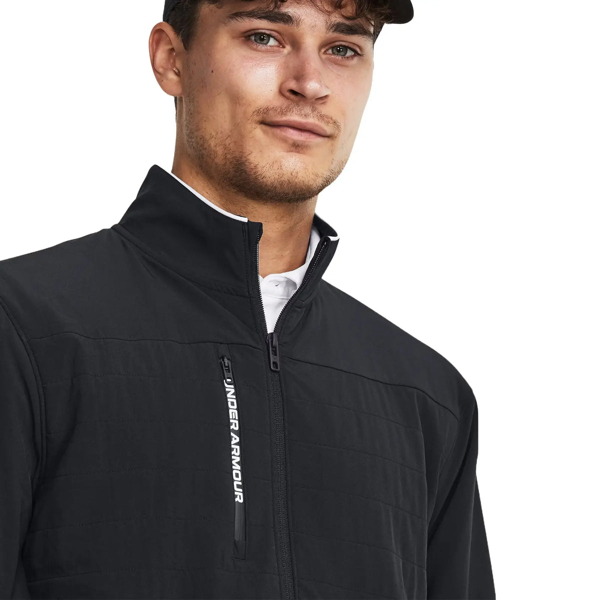 A young man is smiling while wearing a black zip-up jacket with a high collar and a logo on the front. He is in a neutral background.