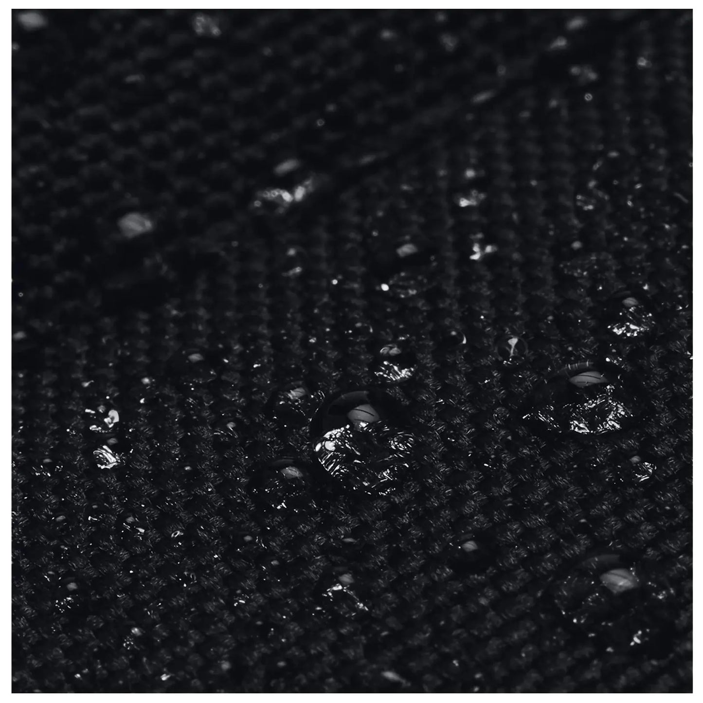 A textured black fabric is covered with small water droplets reflecting light while resting on a flat surface in a potentially dimly lit environment.