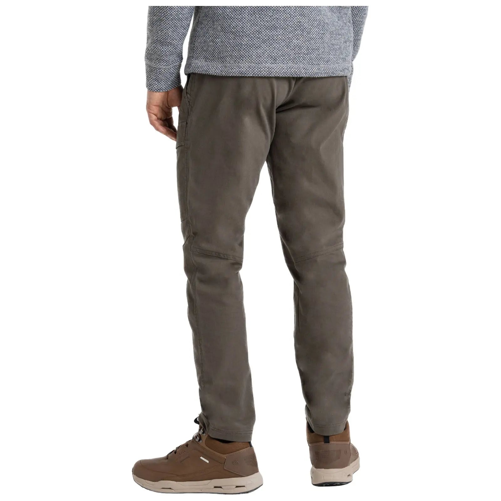 A person stands with their back facing the viewer wearing olive green pants and brown sneakers while dressed in a gray patterned top in a neutral setting.