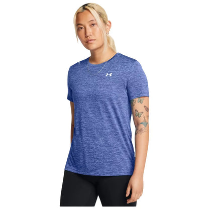 A person wearing a blue athletic shirt with a logo stands confidently in a neutral background showcasing their tattoos and short hair, conveying a casual yet active demeanor.
