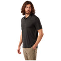 A man with curly hair is wearing a black striped polo shirt and beige trousers while standing with one hand in his pocket and looking to his left against a white background.