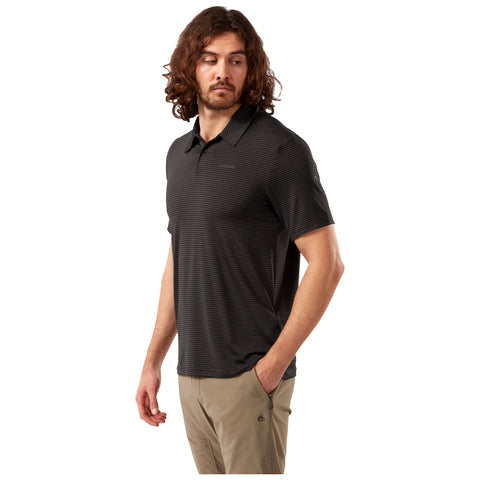 A man with curly hair is wearing a black striped polo shirt and beige trousers while standing with one hand in his pocket and looking to his left against a white background.