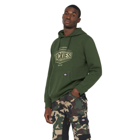 A man stands confidently wearing a green hoodie featuring a logo and camouflage patterned pants, posing against a plain white background.