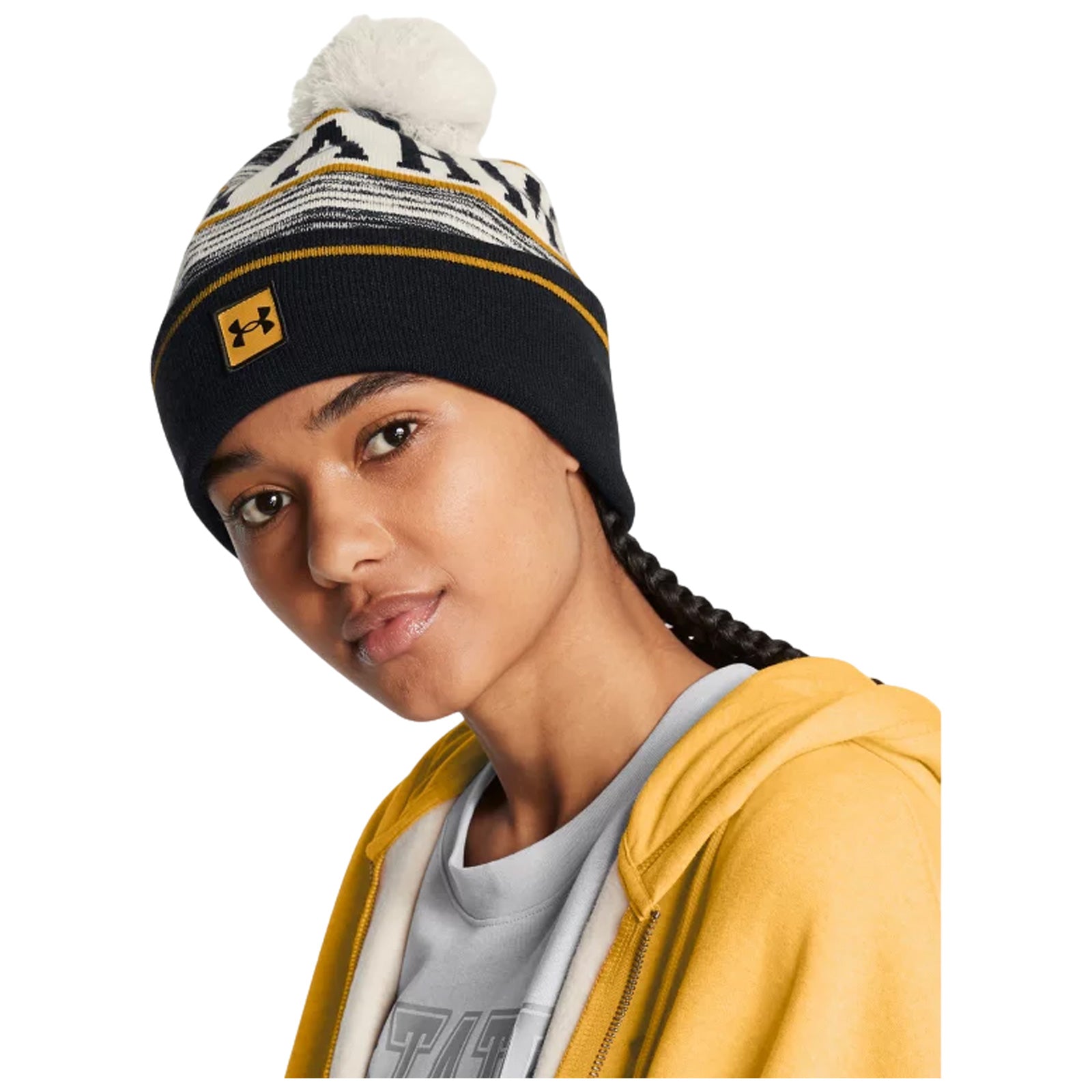 A young woman with braided hair wears a black knitted beanie featuring a pom-pom and a logo while seated in a casual yellow hoodie over a grey shirt, appearing relaxed and engaged.
