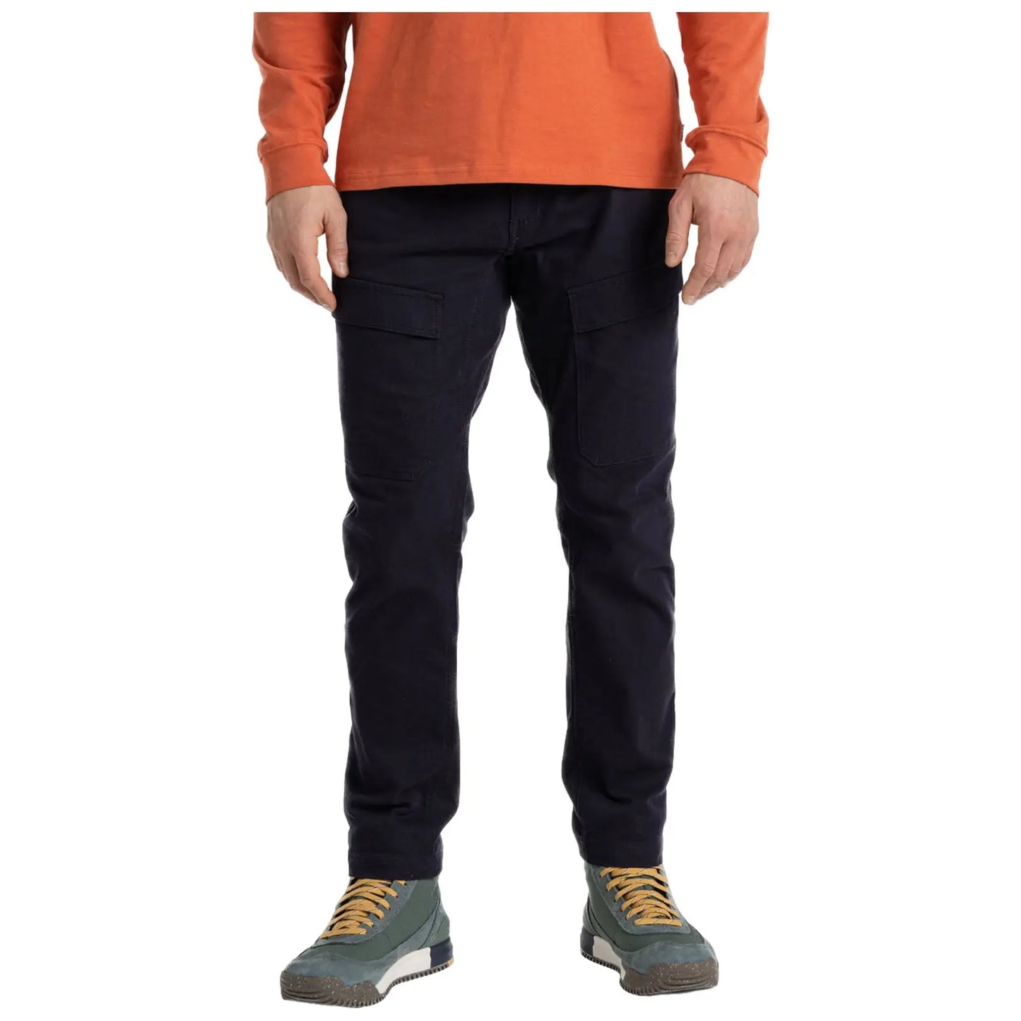 A person wears navy cargo pants and a rust-colored long-sleeve shirt while standing upright. Their footwear is green and grey with yellow laces, set against a neutral background.