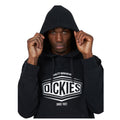 A person wears a black hoodie while pulling the hood over their head with a serious expression in a plain background without distractions. The hoodie displays the text "QUALITY WORKWEAR DICKIES SINCE 1922."