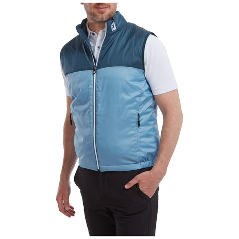 A man is wearing a sleeveless blue and teal vest over a white shirt and black pants while standing with one hand on his hip in a neutral setting.