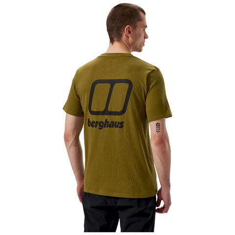 A man wearing an olive green t-shirt with a large black logo on the back stands facing slightly to the right in a plain background