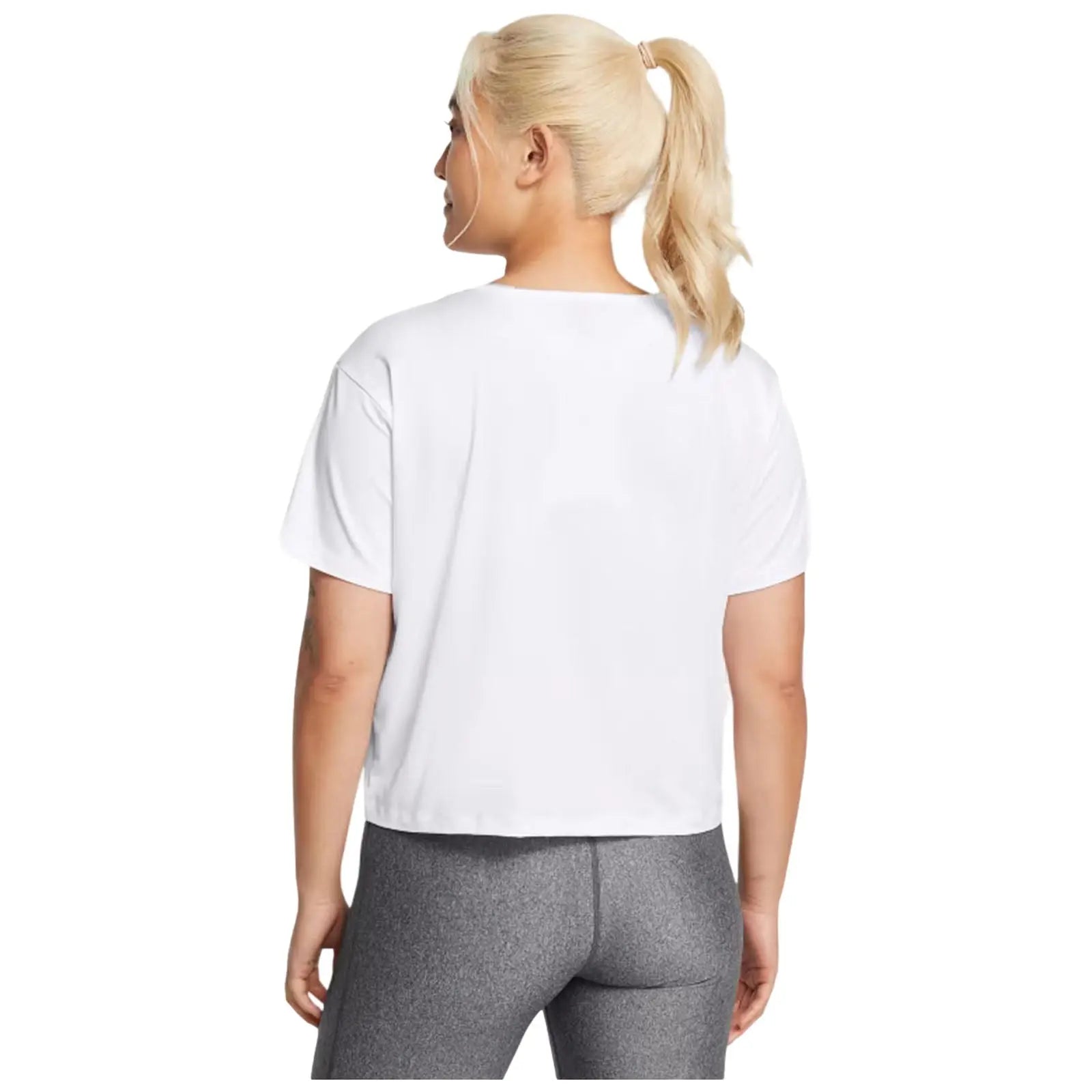A person with light blonde hair tied in a ponytail wears a short white shirt and gray leggings while standing with their back turned in a plain background.