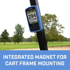 A golf GPS device is mounted on a cart frame displaying hole information and distance measurements in a green golf course environment with trees in the background. Integrated magnet for cart frame mounting.