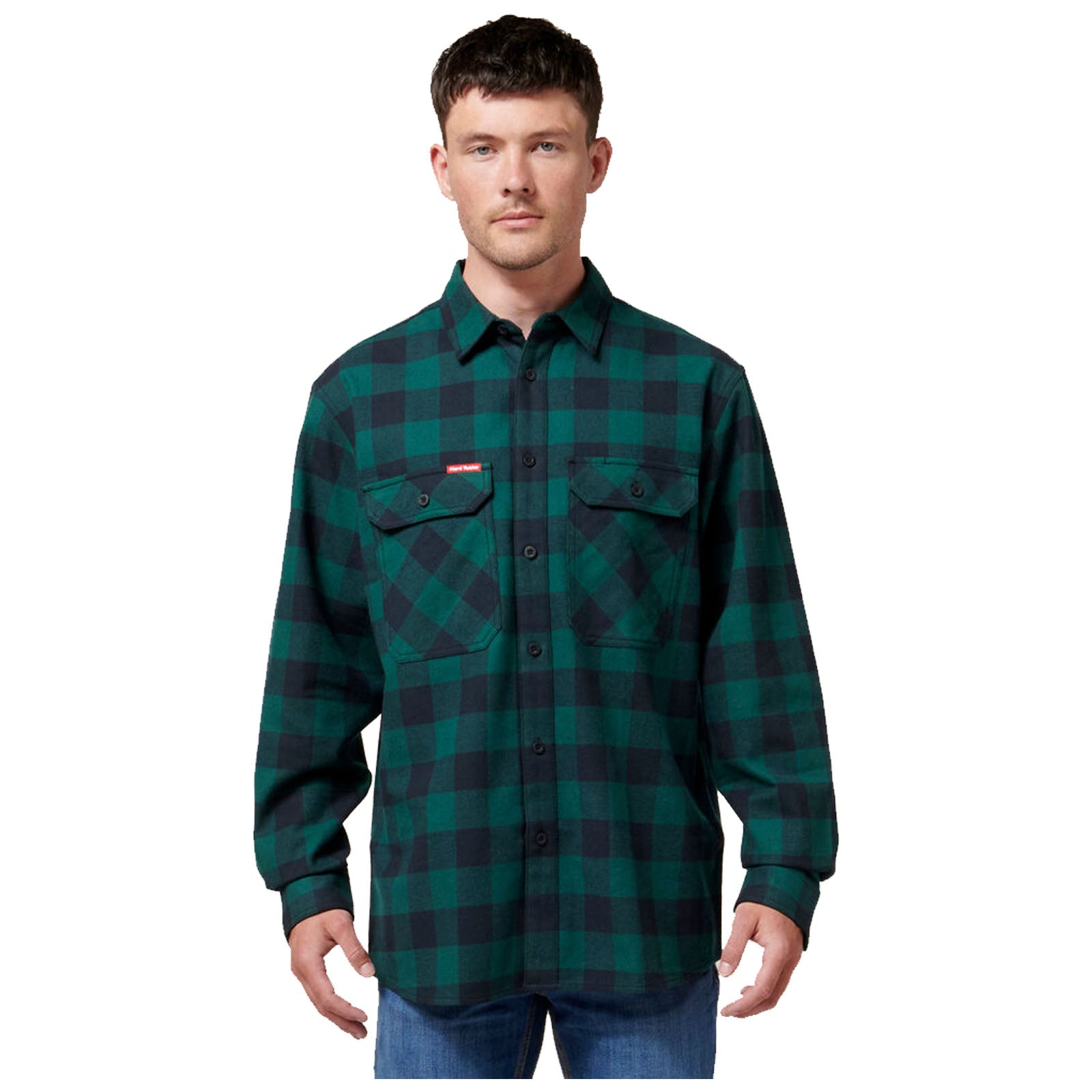 A young man stands confidently wearing a green and black checkered flannel shirt with two chest pockets and a small logo on the left side. He has a relaxed expression.