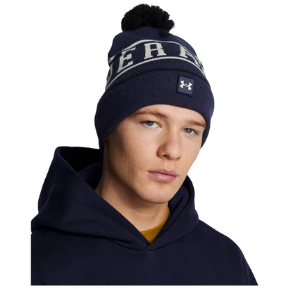A young man is wearing a navy blue beanie with a pom-pom on top and a logo while looking slightly to the side against a plain background, dressed in a dark hoodie.