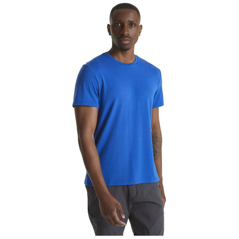 A man stands confidently wearing a bright blue t-shirt and dark pants with a neutral expression in a plain background, suggesting a casual or relaxed setting.