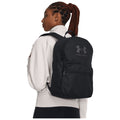 A woman with braided hair wears a black backpack while looking to the side. She is dressed in a light-colored top, standing against a plain background.