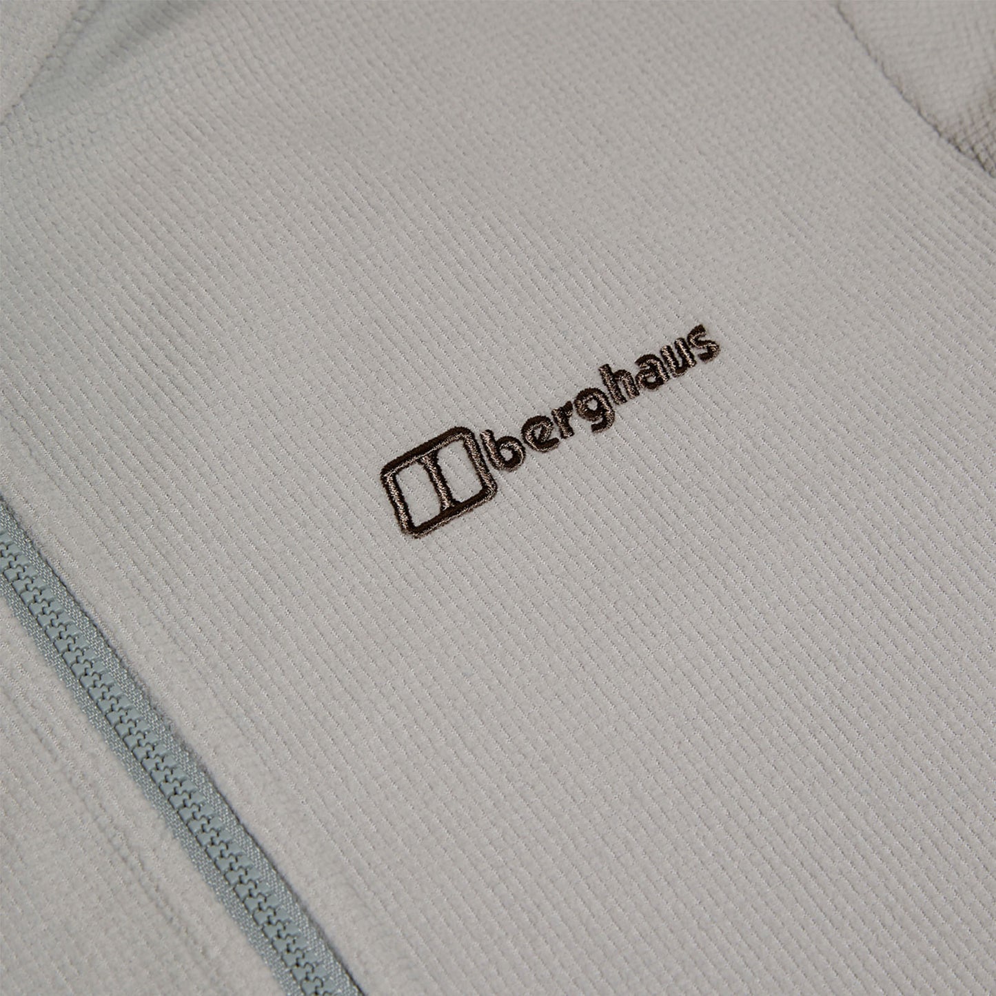 A light-colored fleece garment lies flat displaying a textured fabric with a zipper on one side featuring the embroidered brand name Berghaus in brown near the upper left.