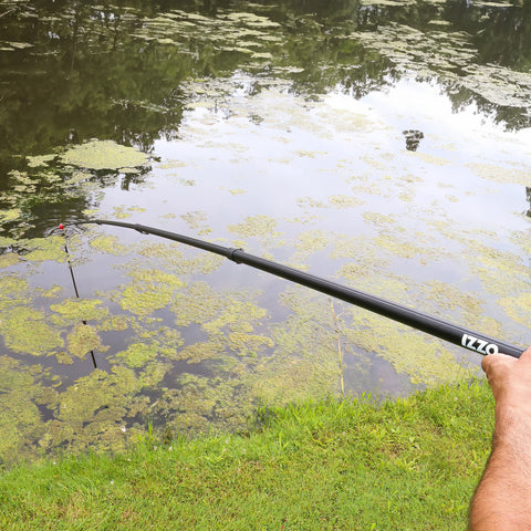 A fishing rod extends over a pond where a person is attempting to catch fish surrounded by water covered in green algae and grass lining the bank under overcast skies.