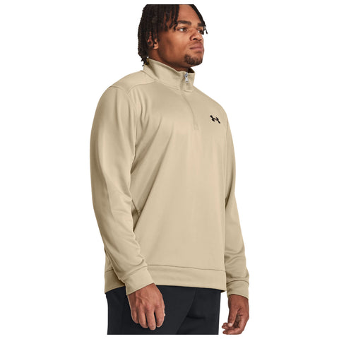 A man is standing confidently wearing a light beige quarter-zip sweatshirt with a black logo on the chest in a neutral background, presenting a casual athletic style.
