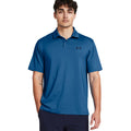 A young man stands confidently wearing a blue polo shirt with a logo on the left chest his hands are casually placed in his pockets in a neutral background.