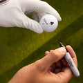 A gloved hand holds a Titleist golf ball while another hand with a marker prepares to write on it in a green outdoor setting.
