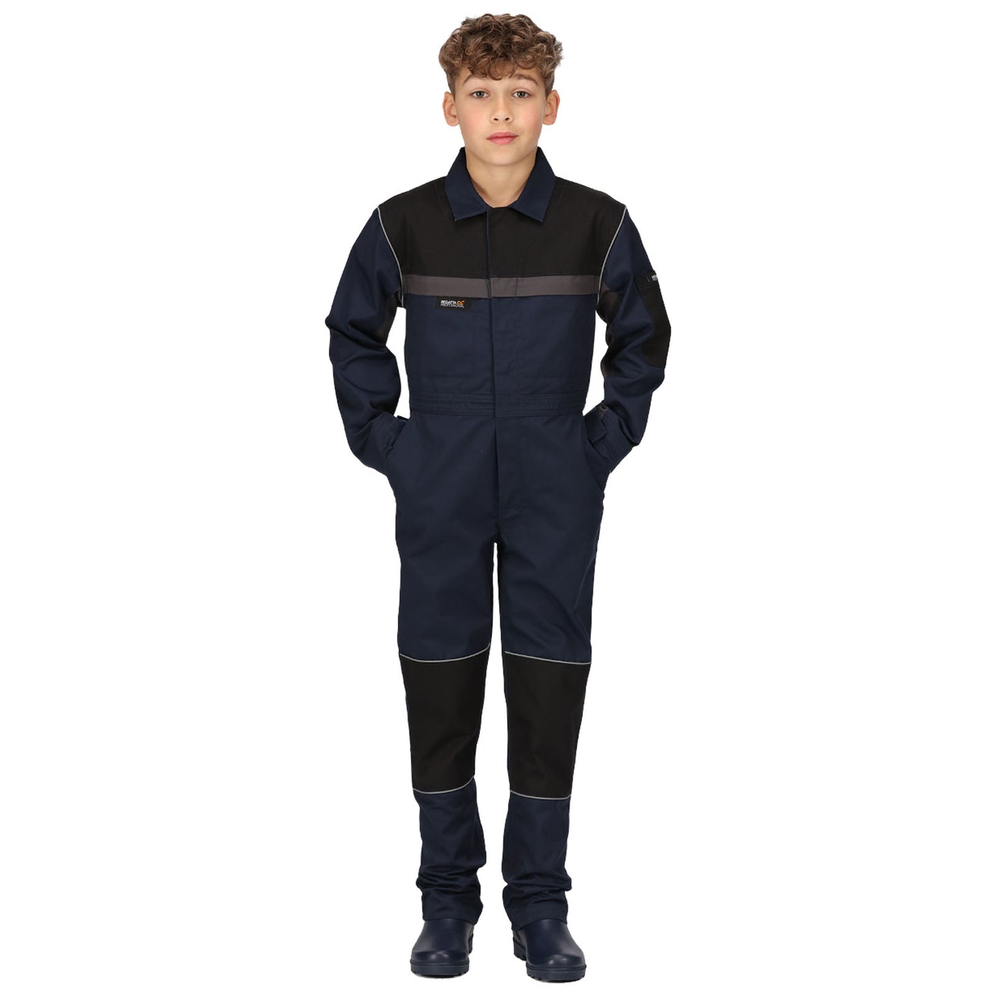 A boy stands confidently in a navy blue jumpsuit with black accents and reflective stripes hands in the pockets on a plain white background suggesting a work or industrial setting.