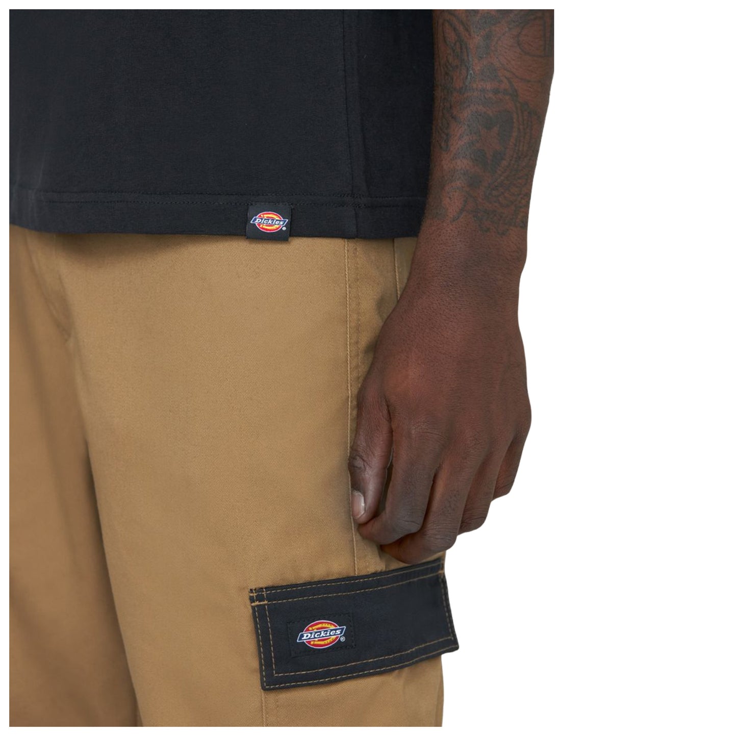 A hand is resting on the side of khaki pants featuring a black patch with the logo of Dickies while wearing a black shirt the scene focuses on casual clothing details.