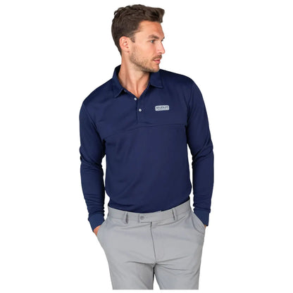 A man stands with his hands in his pockets wearing a long-sleeve navy polo shirt and light gray trousers against a plain white background. He looks to the side.