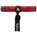 A red paddle with a circular grip is positioned upright while a black strap attached to the handle dangles downward in a neutral background, showcasing its features and branding.