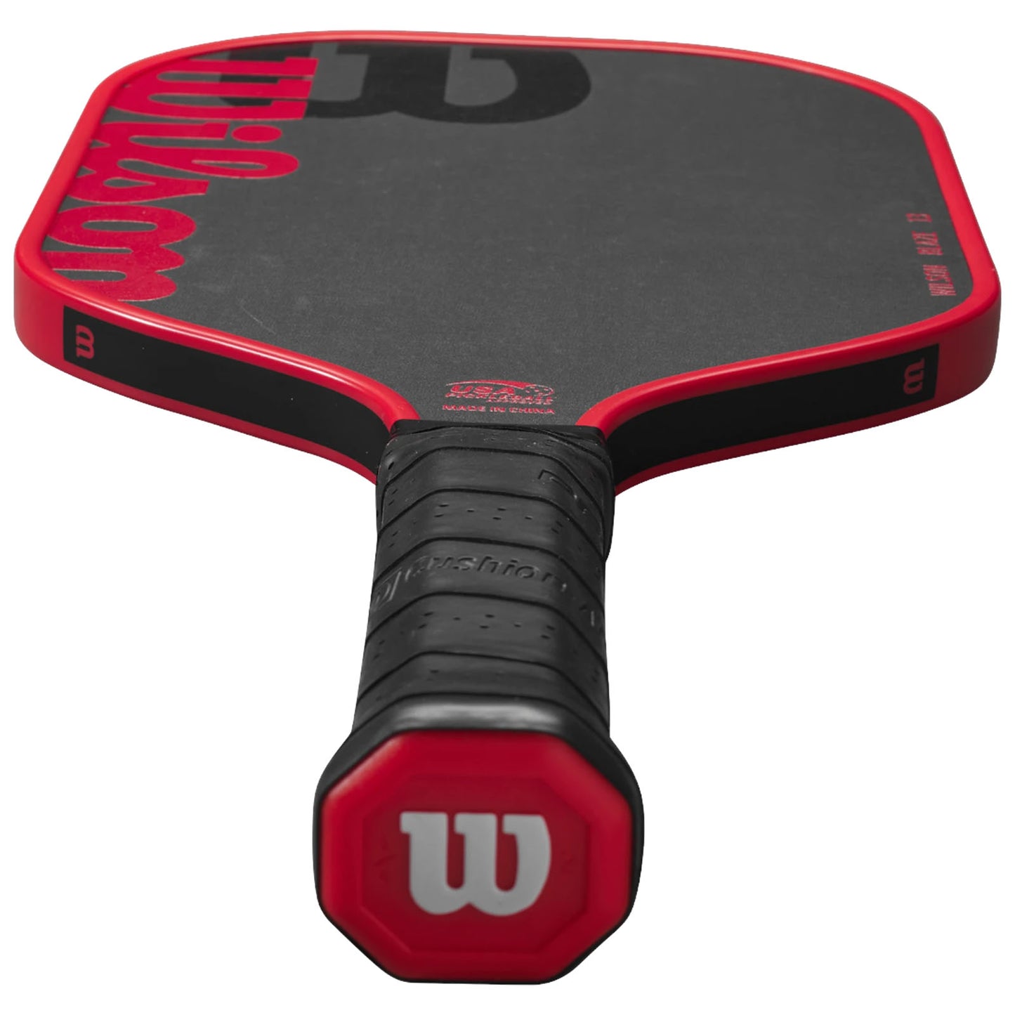 A paddle with a red and black design rests horizontally on a flat surface featuring a textured grip handle and a smooth hitting surface indicating its use in a racquet sport.