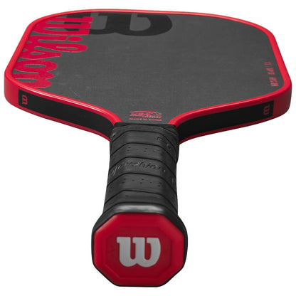 A paddle with a red and black design rests horizontally on a flat surface featuring a textured grip handle and a smooth hitting surface indicating its use in a racquet sport.