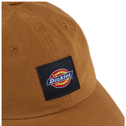 A tan cap with a textured fabric features a black rectangular patch displaying the "Dickies" logo in colorful lettering, placed against a neutral background.