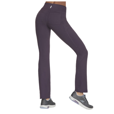 A person wearing dark purple leggings stands with one leg slightly lifted showcasing the fit and style of the pants paired with gray athletic shoes in a neutral background.