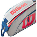 A gray tennis bag with red and blue accents is partially unzipped revealing its storage compartment the bag features a prominent Wilson logo on the side and a carrying strap.