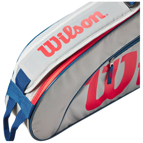 A gray tennis bag with red and blue accents is partially unzipped revealing its storage compartment the bag features a prominent Wilson logo on the side and a carrying strap.