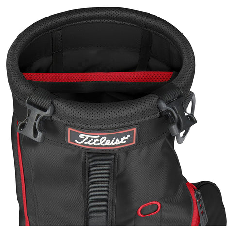 A black golf bag with a round top and a red interior features the Titleist logo and a buckle for securing the top. The bag displays various storage compartments and straps.