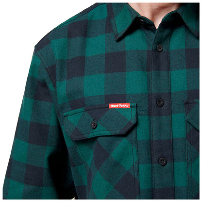 A green and black plaid shirt features two chest pockets with buttons and a label that reads Hard Yakka positioned on the upper left side of the shirt.