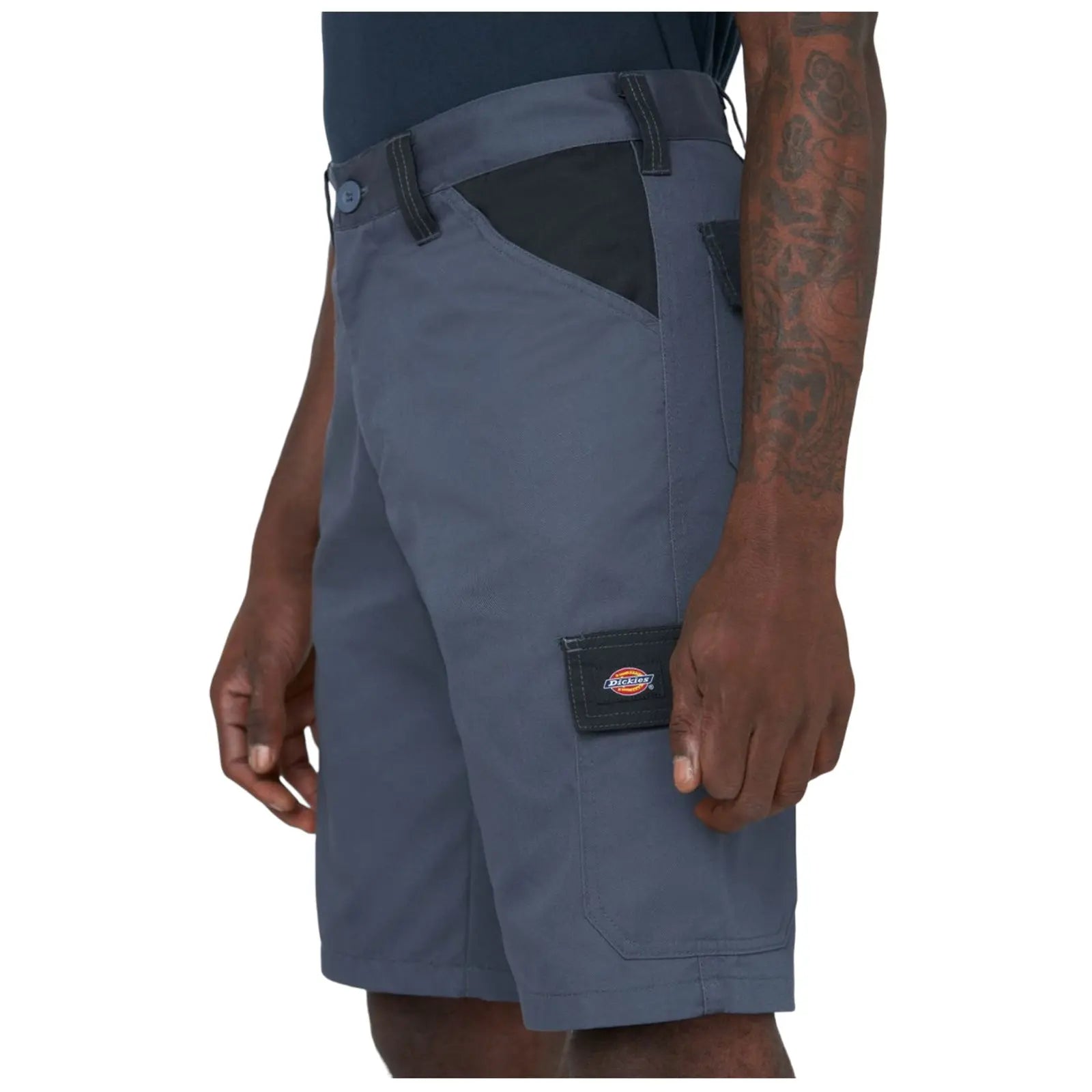 Gray cargo shorts are being worn by a person who stands with one hand near the pocket displaying a logo on the side in a neutral background setting.