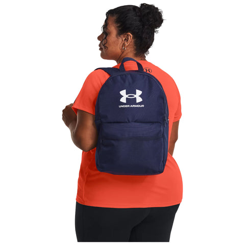 A woman stands facing slightly away while wearing an orange shirt and a navy blue backpack with a white logo on it in a neutral setting.