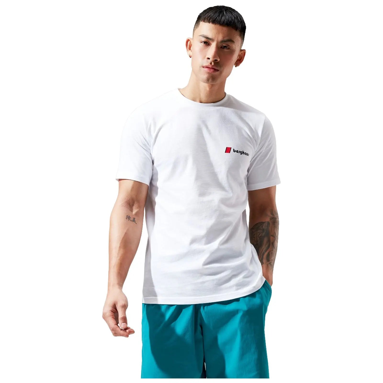 A man stands in a white t-shirt and turquoise shorts with a confident expression his right arm raised slightly displaying a tattoo in a minimalistic, bright background.