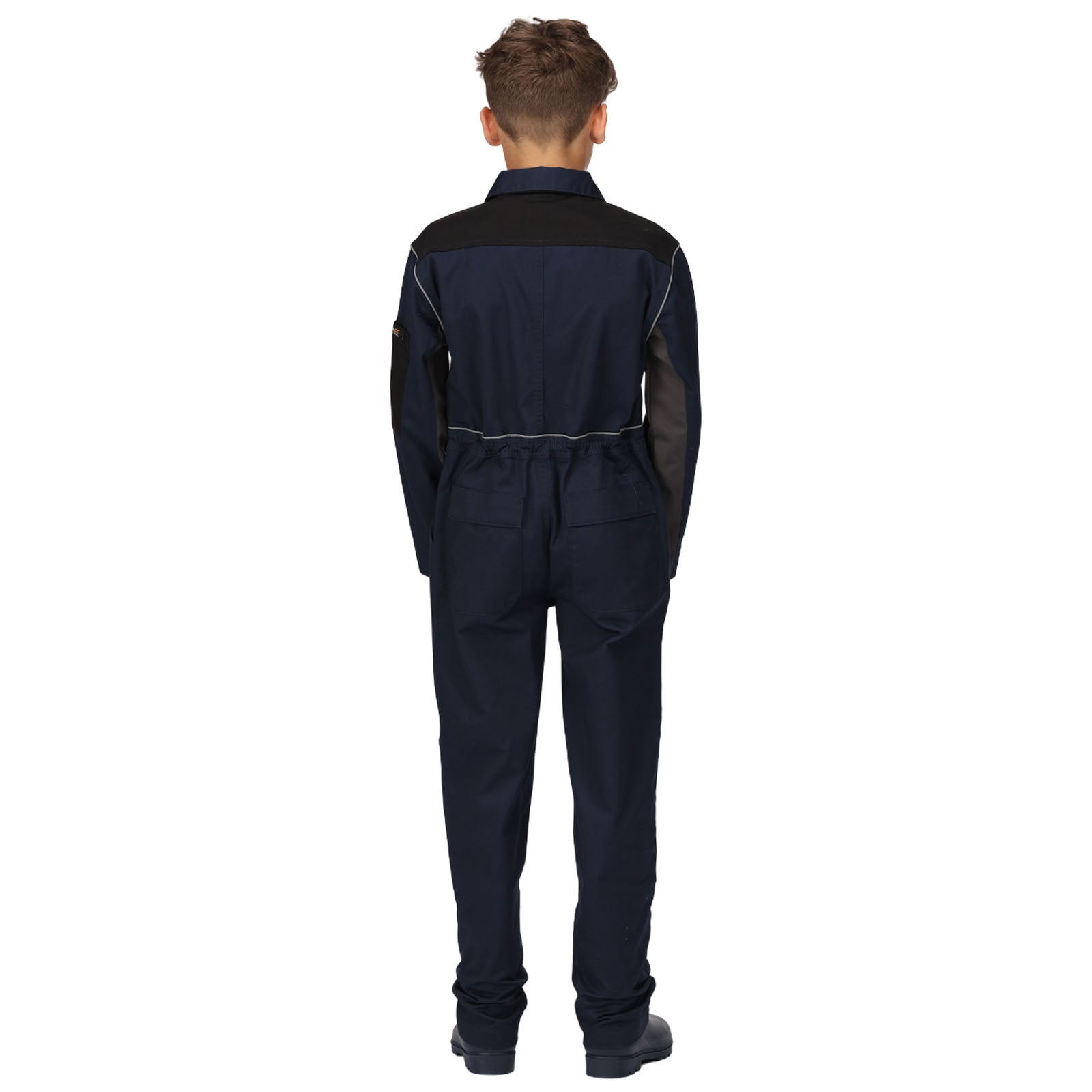 A child wears a navy blue jumpsuit with black accents standing with their back facing the viewer in a minimalistic setting highlighting the design of the outfit.