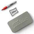 A Titleist silver tin box rests flat with a black and white Titleist logo pin beside a red and gray marker labeled Titleist on a white background.