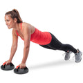 A woman in a red athletic top performs push-ups using push-up handles on a plain white background while maintaining a focused expression and proper form.