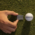 A hand holds a gray golf tee while positioning it next to a white golf ball on green grass preparing for a shot in a golfing context.